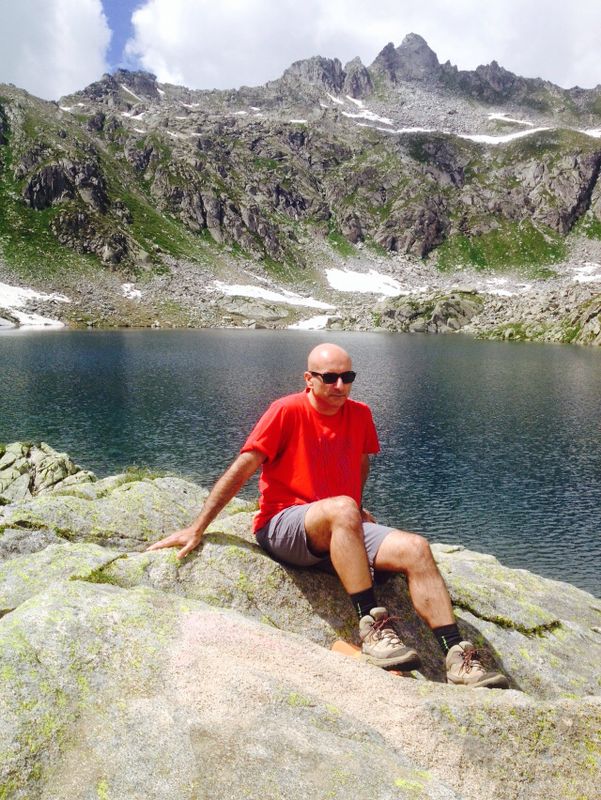 Dr. Ugo Cavallaro wearing sunglasses, sitting on rock next to water and mountains