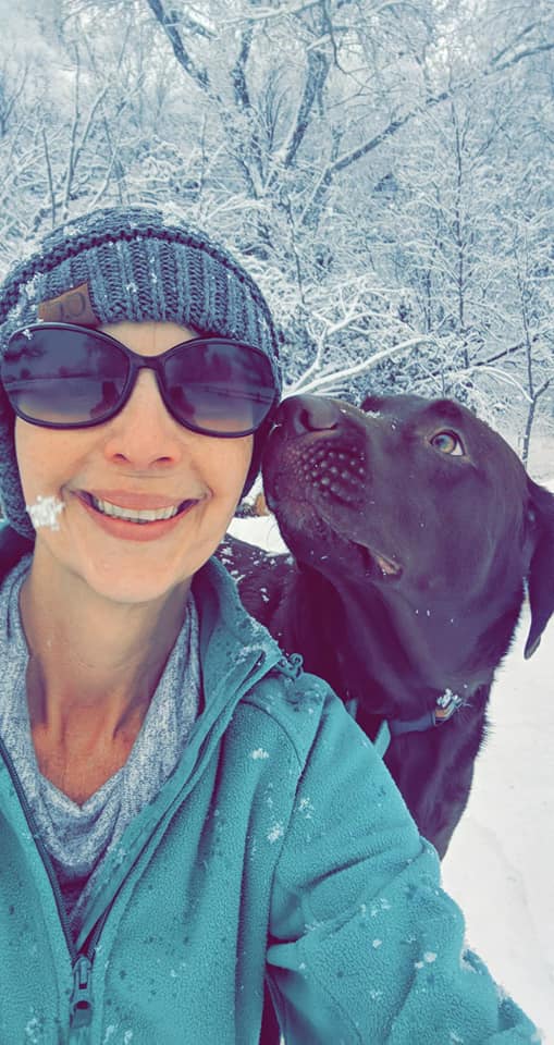 Stacy Saravo, wearing a winter hat and posing for a selfie in the snow as her dog gives her a kiss 
