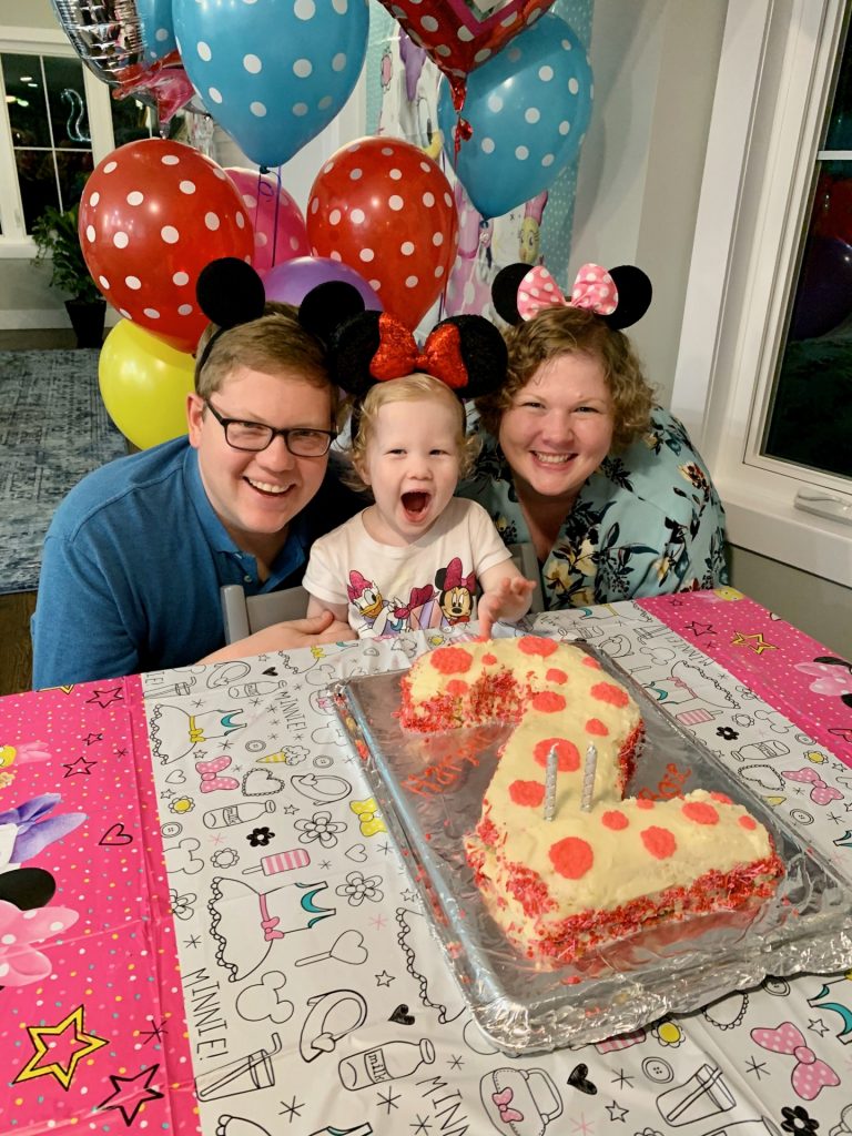 Jess with husband and daughter, all wearing Micky and Minnie Mouse ears, in front of balloons and cake shaped like the number 2