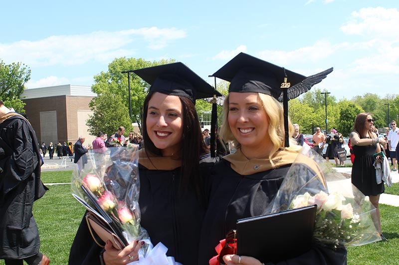 Morgan Gaynor with friend, both wearing cap and gown and holding flowers and diploma