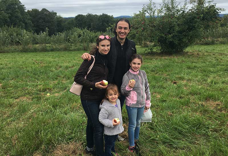 Photo: Dr. Dmitri Zamarin, smiling broadly as he stands in a lush green field with a woman and two children, all holding apples.