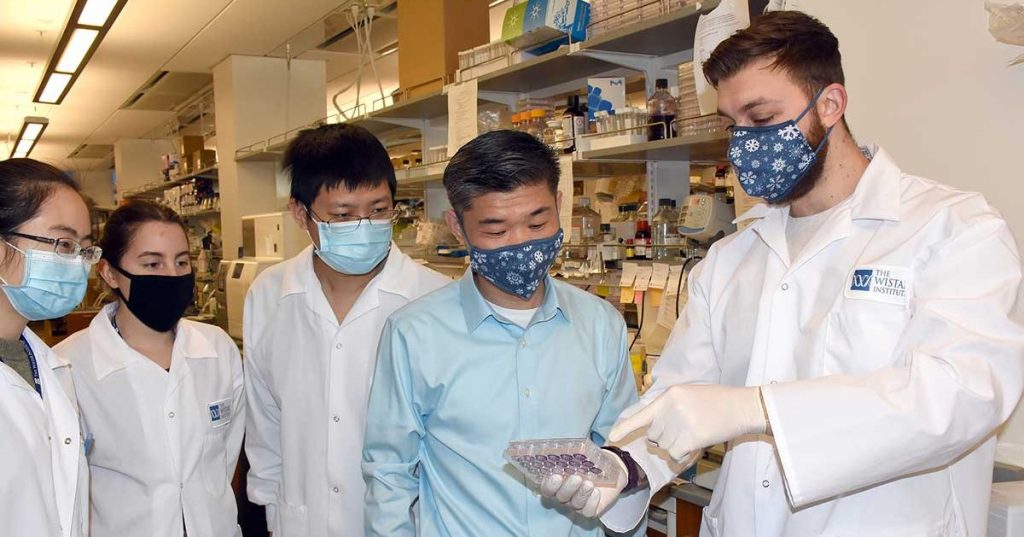 Rugang Zhang, PhD, masked with four members of research team in lab, looking at specimen