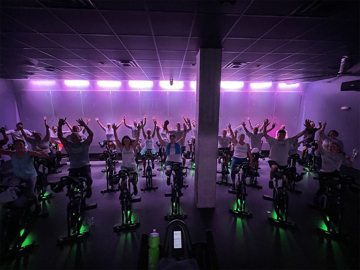 large group of indoor cycling riders in purple lit studio, on stationary bikes, with arms in in victory signs