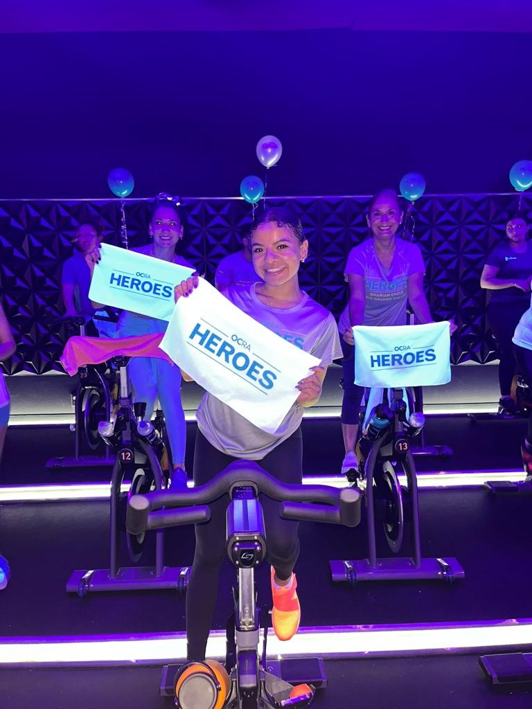 Young women on stationary bikes in fitness class, smiling and holding signs that say OCRA Heroes