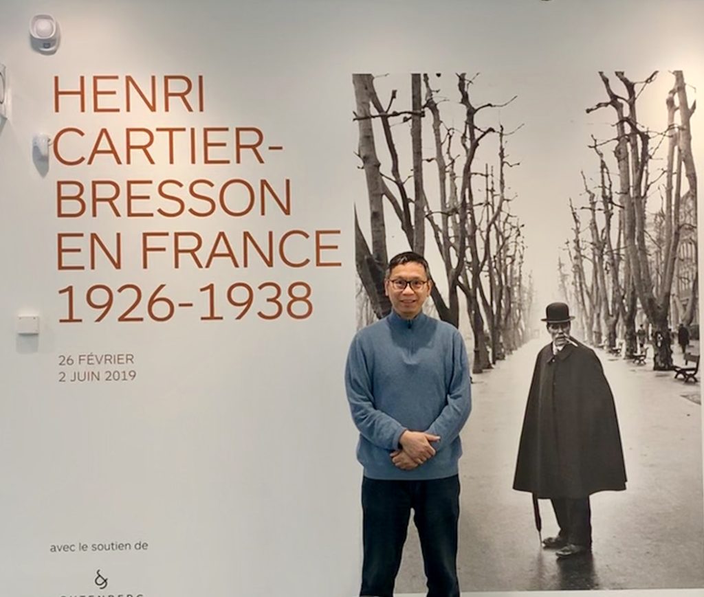 Ie-Ming Shih posing in art gallery in front of piece with text Henri Cartier-Bresson En France 1926-1938