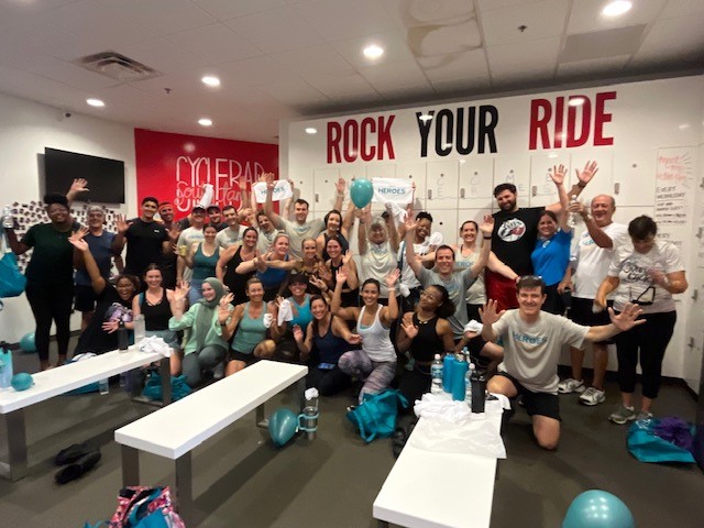 group of people in gym with wall that reads Rock Your Ride, holding arms up and smiling