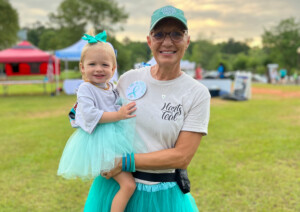 Photo: Smiling woman wearing a teal baseball cap and teal tutu, standing at an outdoor event. She's holding a smiling little girl who is wearing a teal bow in her hair, and teal tutu. 