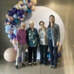 Marsha posing with a group of three other women, all standing next to a backdrop of multicolored balloons
