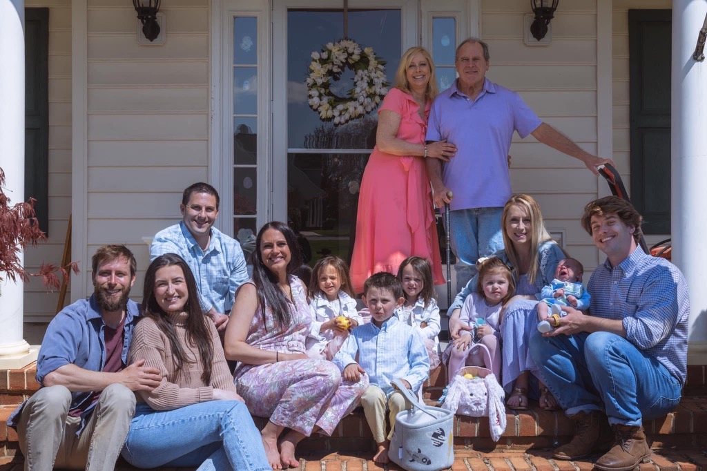 Janice standing in a pink dress, with her husband, posing with their large family on the porch.