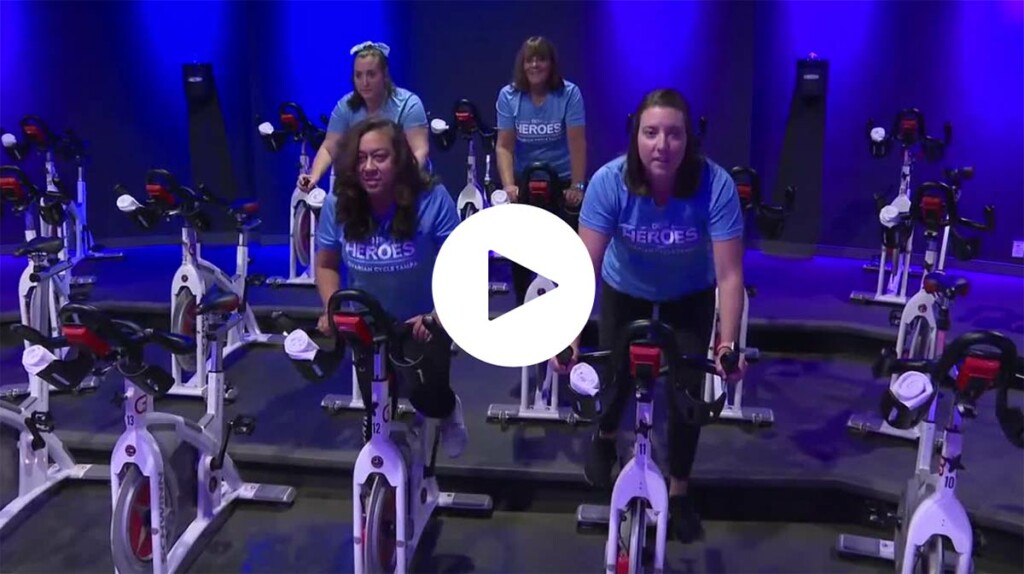 Still image of video from news broadcast, showing people in a dark purple fitness studio, on stationary bikes. A play button appears in the middle of the still.