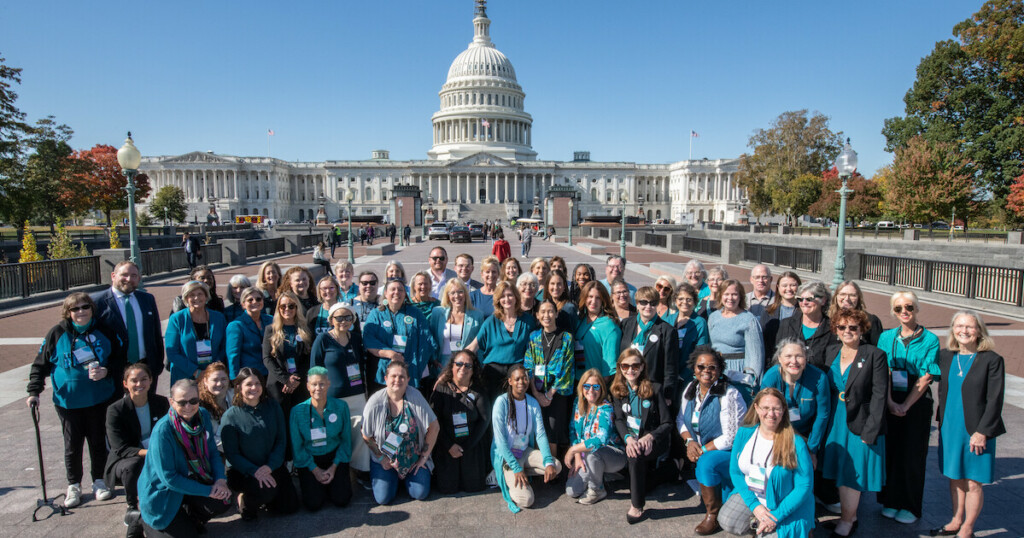 A large diverse group, all wearing teal, kneels and stands in front of the Capitol building. All are wearing buttons that read OCRA.