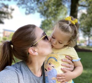 A woman wearing sunglasses and a gray shirt kisses a small child on the lips. The child, dressed in a yellow shirt with a Disney character and a yellow bow in her hair, looks content. They are outdoors with trees and greenery in the background.