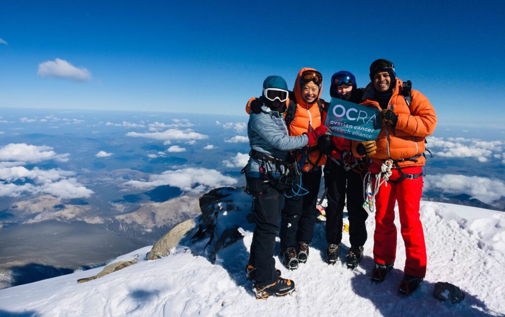 Four climbers in winter gear stand at a snowy summit, celebrating their ascent. Three individuals hold a blue banner that reads "OCRA Ovarian Cancer Research Alliance." The background shows a panoramic view of snow-capped peaks and a clear, blue sky.