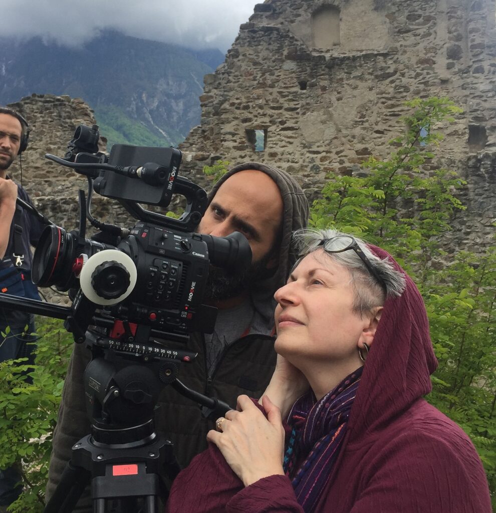 Two people are operating a professional video camera in an outdoor setting with ancient ruins and a mountainous backdrop. One person in a hooded jacket is looking through the viewfinder, while the other, wearing glasses, stands beside them, observing.