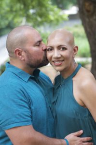 A bald man gently kisses his wife, who is also bald from chemotherapy, on her head as they stand together outdoors. Both wear teal tops, with trees and greenery in the background, creating a warm and affectionate atmosphere.