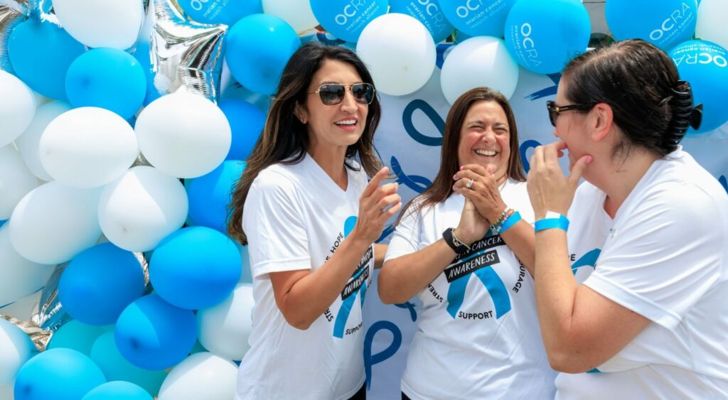 Three women laughing and smiling while standing in front of a backdrop decorated with teal and white balloons. They are wearing white t-shirts with a teal ribbon design, indicating participation in an ovarian cancer awareness event.