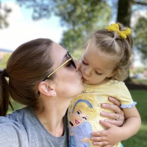 A woman wearing sunglasses and a gray shirt kisses a small child on the lips. The child, dressed in a yellow shirt with a Disney character and a yellow bow in her hair, looks content. They are outdoors with trees and greenery in the background.