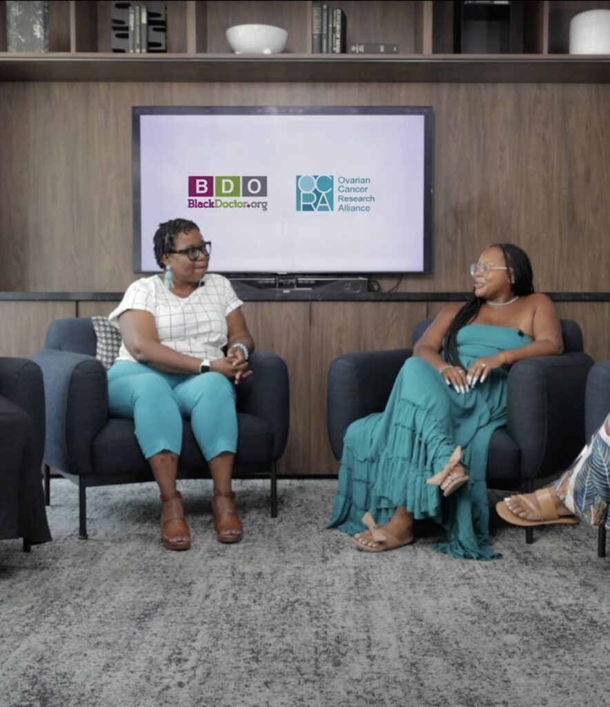 Two women sit in chairs participating in roundtable discussion. Behind them a television screen displays the ocra logo and black doctors logo