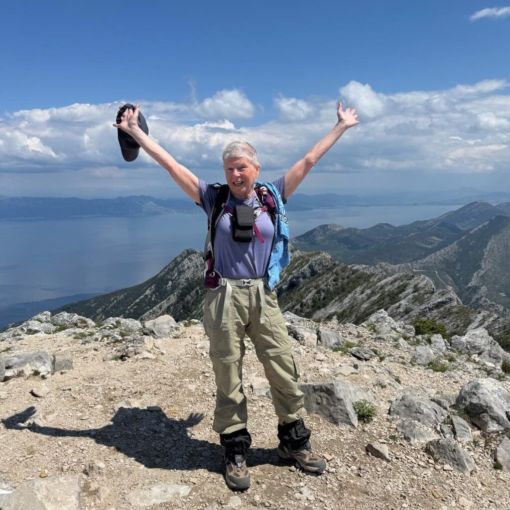 An ovarian cancer survivor wearing hiking gear stands on top of a mountain in front of a blue sky, holding her arms out and looking victorious