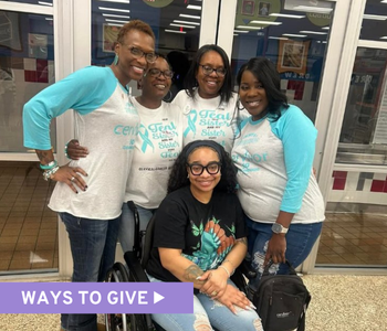A group of four women, wearing matching teal and white shirts with a teal ovarian cancer ribbon, stand smiling with a woman using a wheelchair, who is also wearing teal