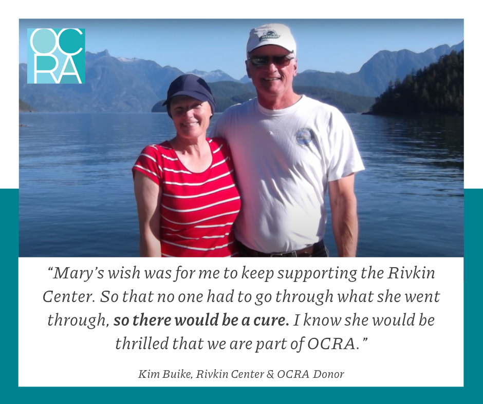 Photo with text overlay. Mary and Kim Buike stand smiling for the camera in front of a gorgeous lake setting. Both are wearing baseball caps. Kim's arm is around Mary. Text reads, "Mary's wish was for me to keep supporting the Rivkin Center. So that no one had to go through what she went through, so there would be a cure. I know she would be thrilled that we are part of OCRA." Kim Buike, Rivkin Center & OCRA Donor