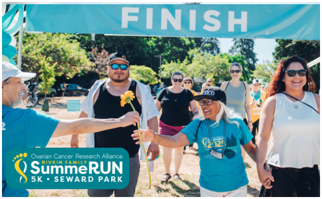 SummeRun participants happily  cross the finish line. A smiling woman accepts a long-stemmed flower from a woman in a teal shirt, as she crosses the finish line with friends. 